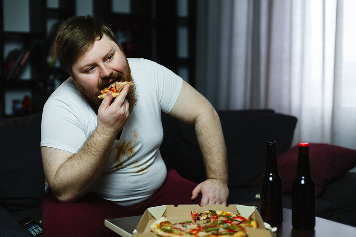 Otyły mężczyzna jedzący pizzę - Przyczyny ginekomastii, czyli jak do tego doszło