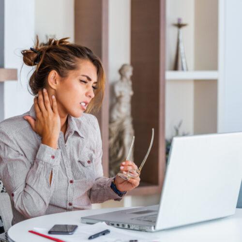 Kobieta przed laptopem dotyka bolącej szyi. Siedzący tryb życia - to nie tylko problemy z krążeniem