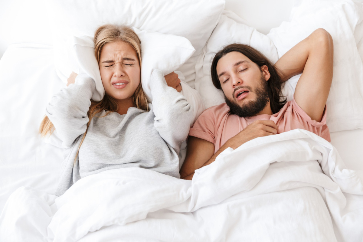 Chrapiący mężczyzna i kobieta zatykająca uszy poduszką. Objawy bezdechu sennego - jak rozpoznać to zaburzenie snu