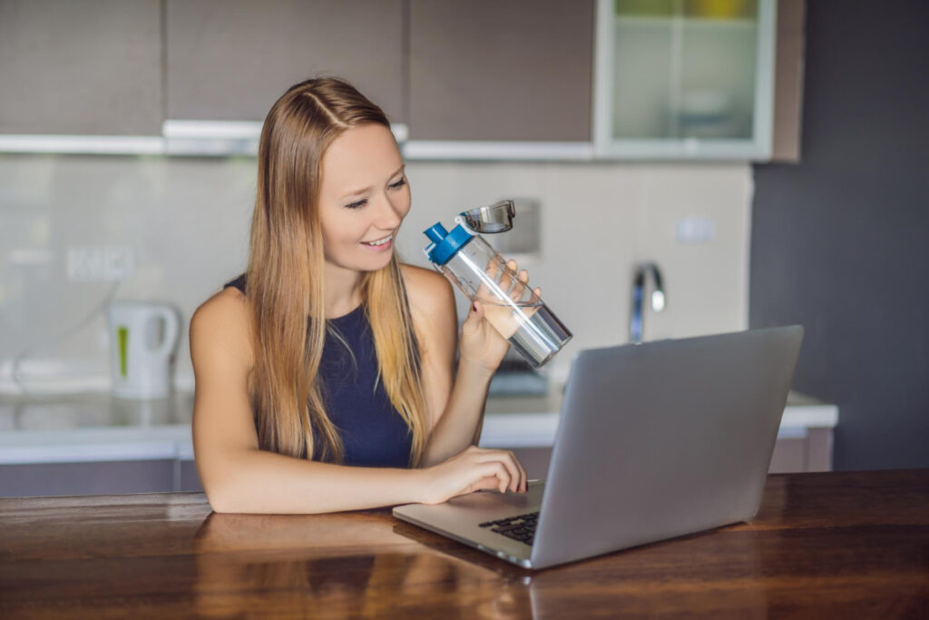 Kobieta pije wodę pracując przy laptopie - picie wody codziennie